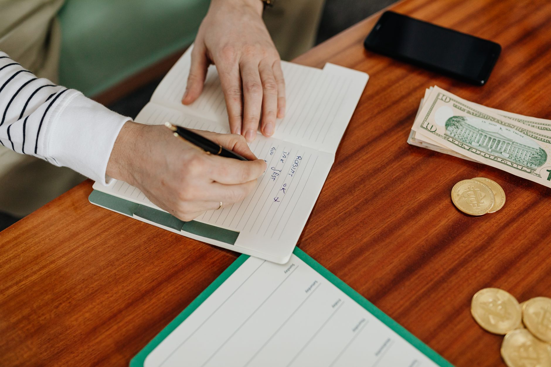 A person writing a household savings plan beside cash, coins, and a phone